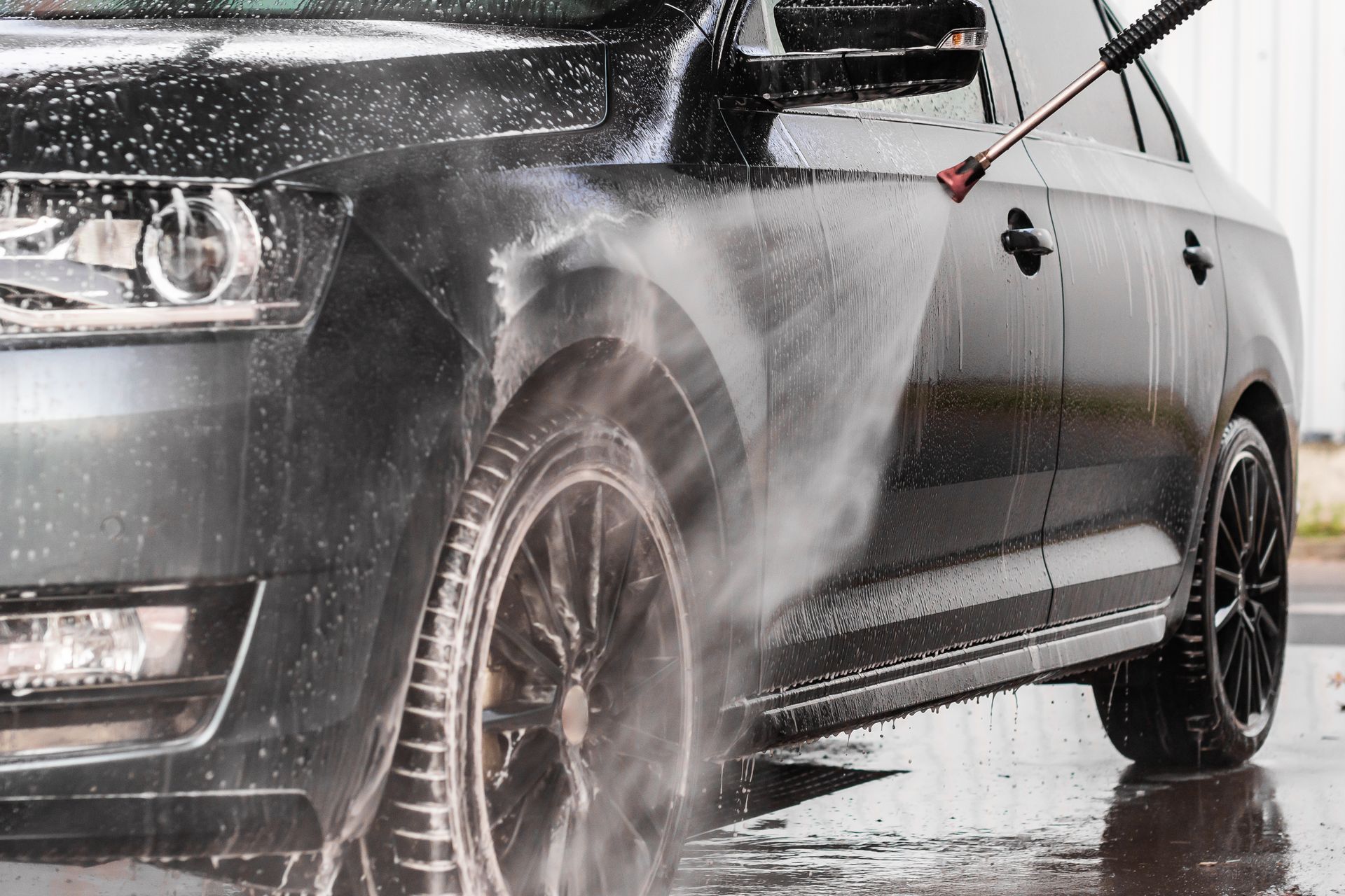 Person washing a black car with a high pressure sprayer, rinsing soap from the exterior.