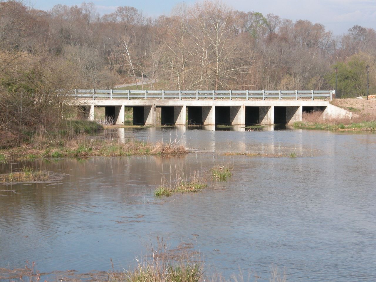 Concrete bridge with rectangular openings over a calm body of water; trees in the background.