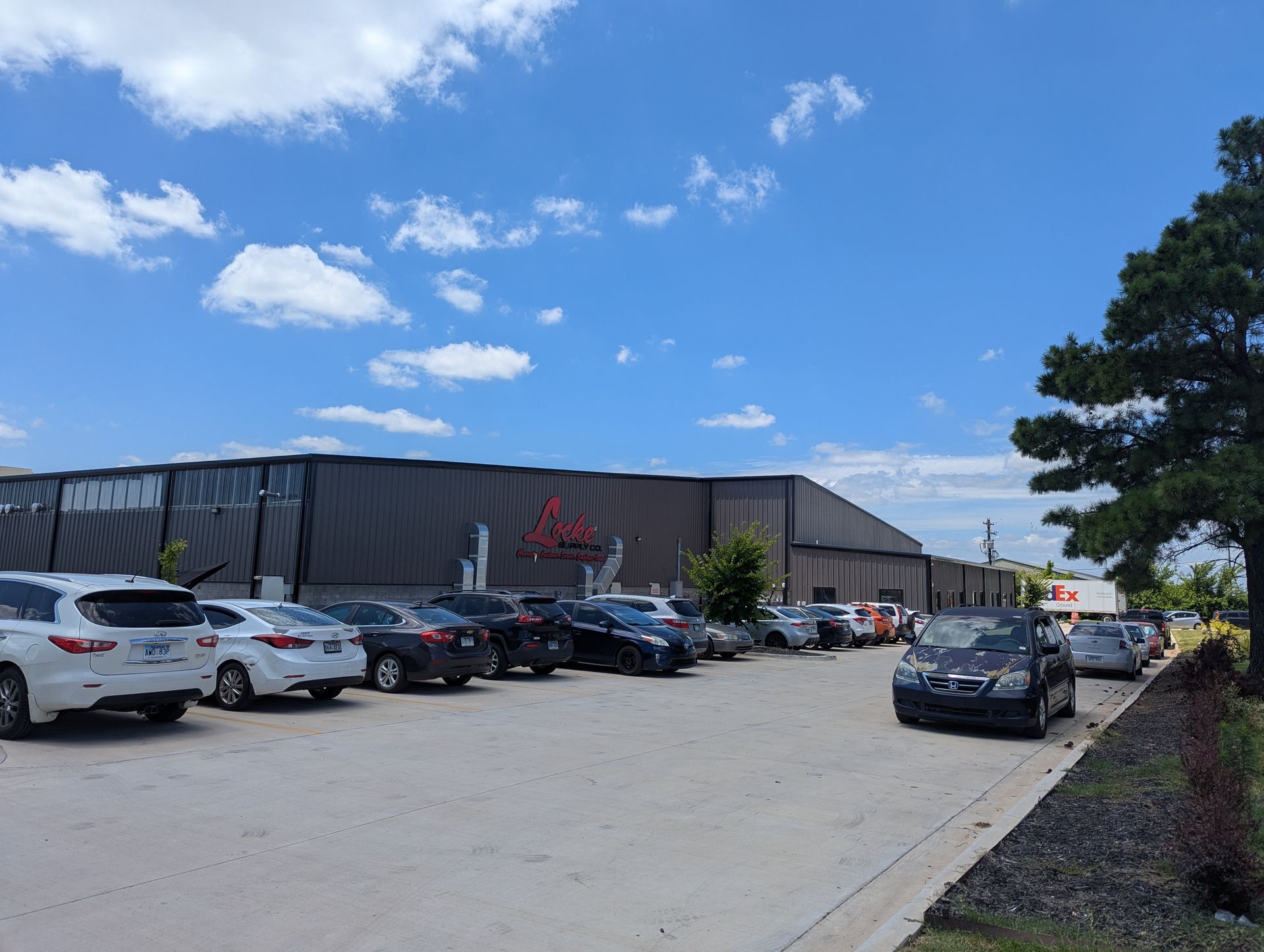 Parking lot with cars in front of a large black building with red logo under a cloudy blue sky.