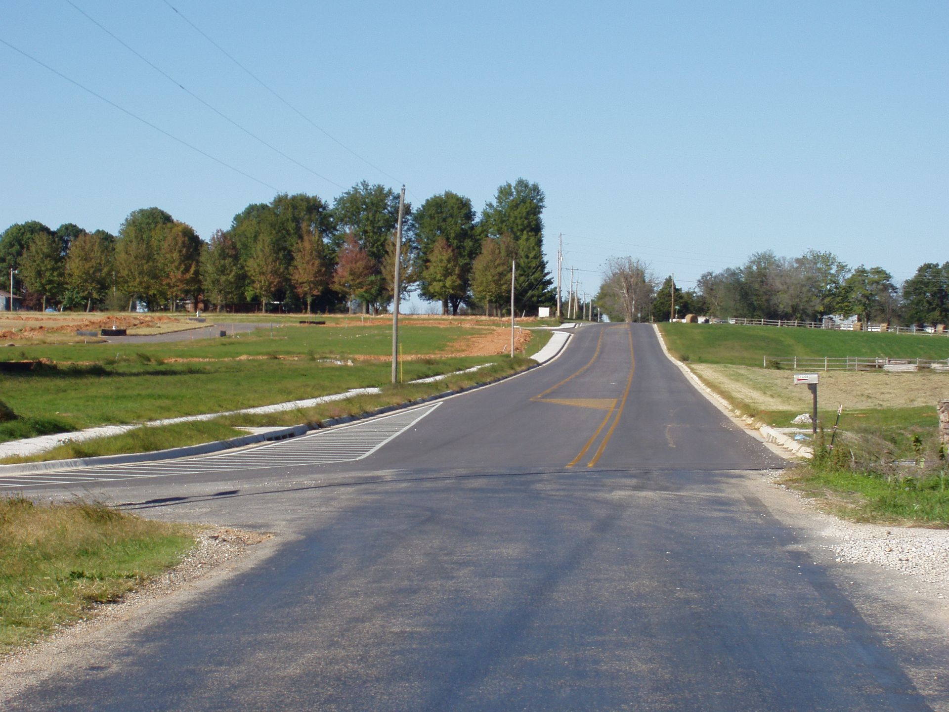 Paved country road ascending a hill under a clear blue sky, bordered by green fields and trees.