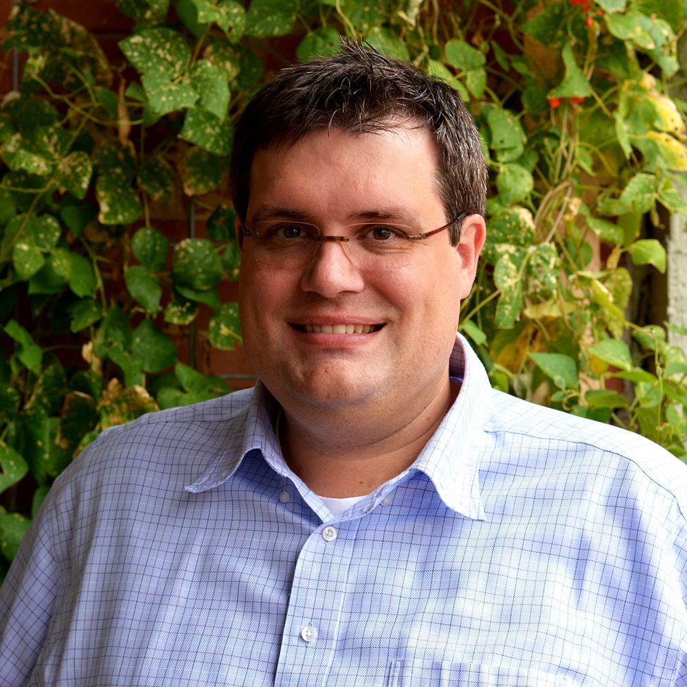 Man with glasses smiling, wearing a blue patterned shirt, set in front of green vines.