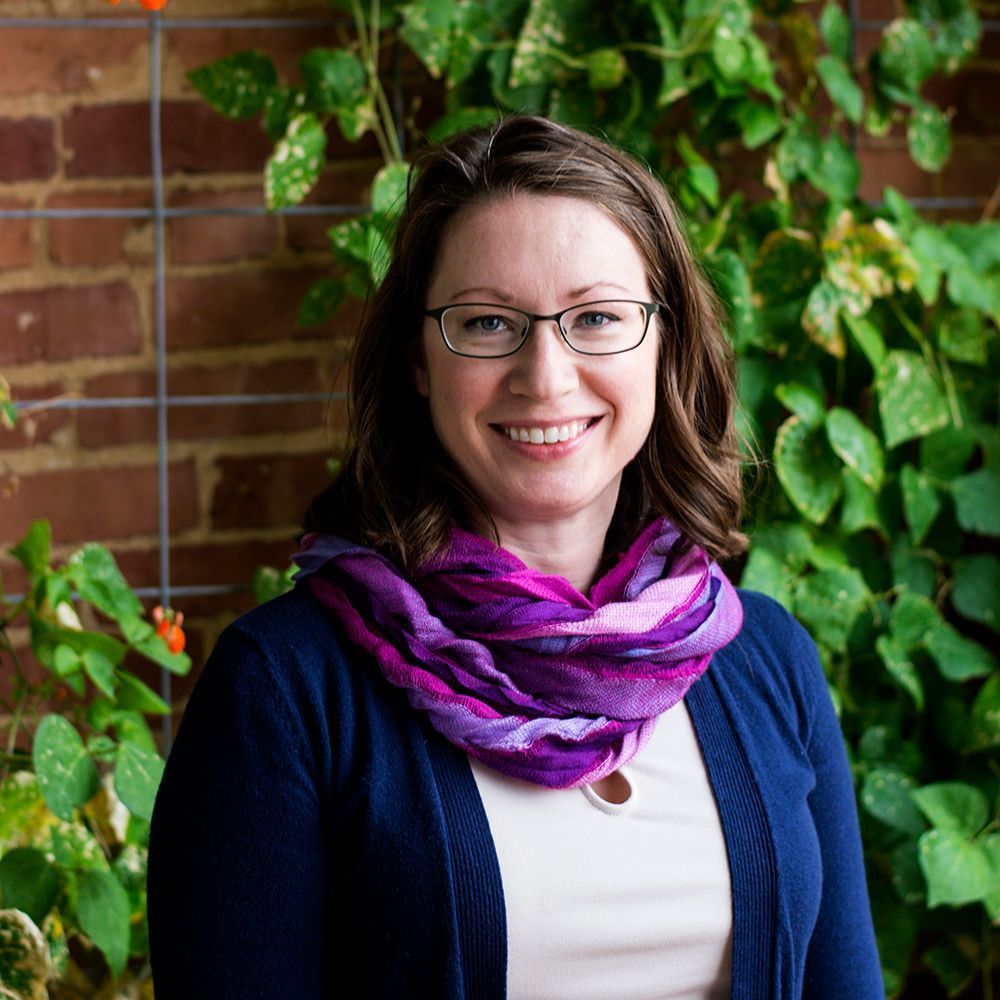 Woman wearing glasses and a purple scarf, smiling, standing in front of vines and a brick wall.