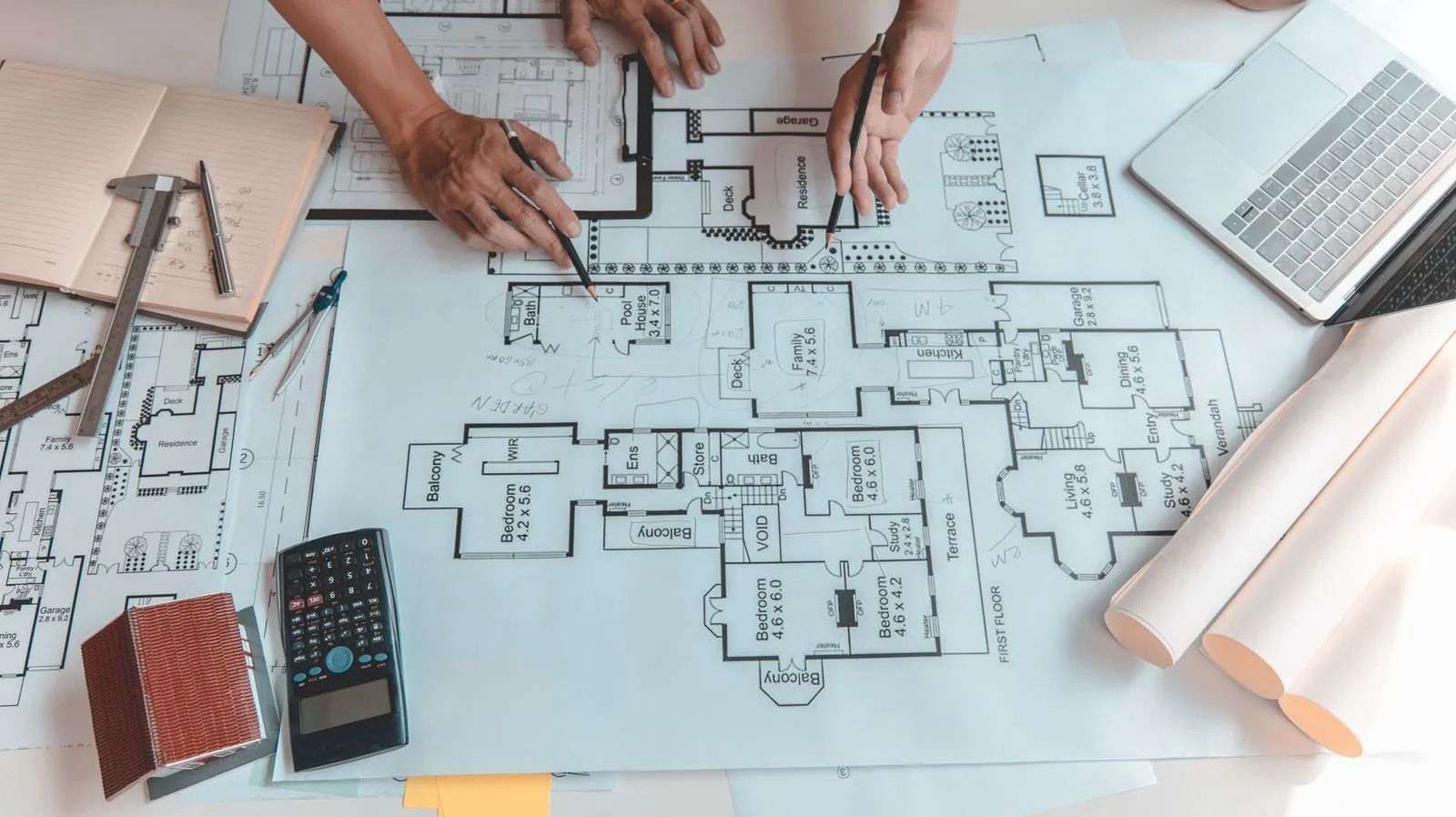 Hands reviewing a floor plan blueprint with pencils, calculator, and laptop on a desk