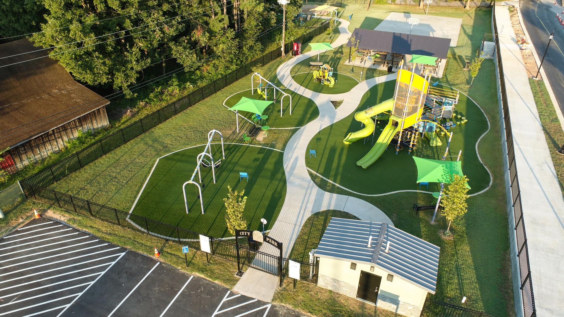 Aerial view of a playground with various play structures, green turf, winding pathways, and a small building.