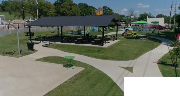 Park with a black-roofed pavilion, picnic tables, playground, and paved paths. Green grass and trees surround the area.