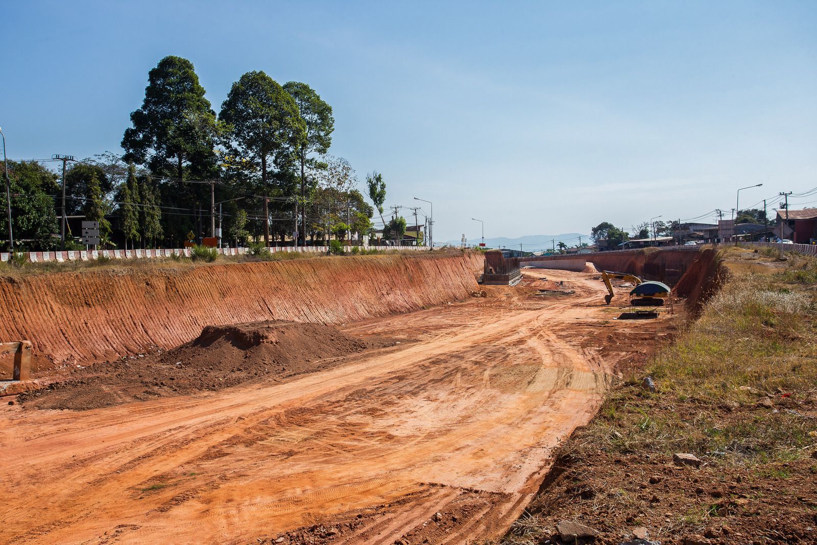 Construction site with dirt road, earth banks, and trees under a blue sky.