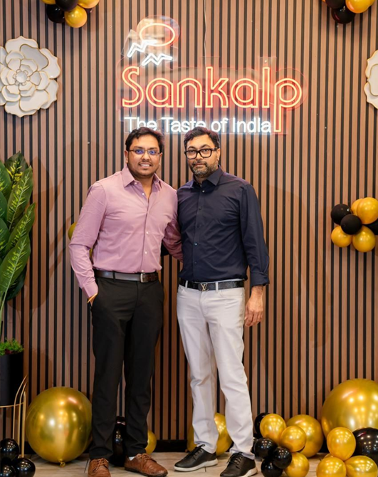 Two men pose in front of a restaurant sign; one wears a pink shirt, the other a blue shirt. Balloons and decorations surround them.