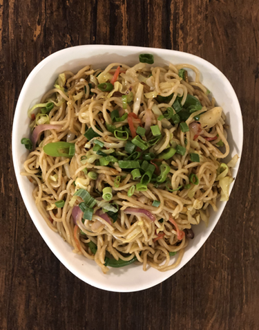 Bowl of noodles with vegetables and green onions on a wooden table.