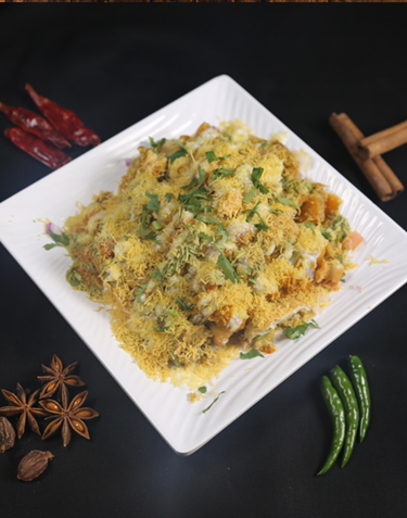 A plate of savory Indian chaat, topped with sev, herbs, and spices, with chili peppers and star anise nearby.