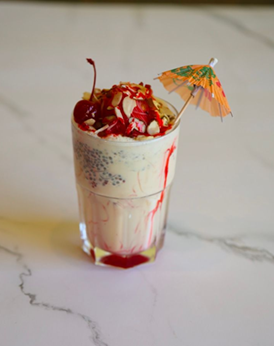 Tall glass of Falooda dessert, topped with cherries, nuts, and a small parasol on a marble surface.
