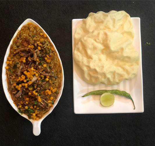 A leaf-shaped bowl of cooked dish with fried onions, papadum, lime, and chili on a white plate.