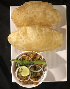 Two puffed-up fried breads (puri) atop a plate with chickpea curry (chole), garnished with onions, lemon, and green chili.
