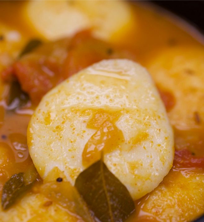 Close-up of a South Indian dish. White, round dumplings in a tomato-based curry, with green curry leaves.