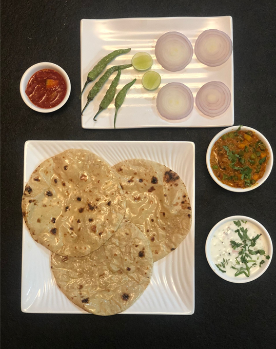 Indian meal: roti, curry, chutney, raita, onions, chilies, and lime slices on a black surface.