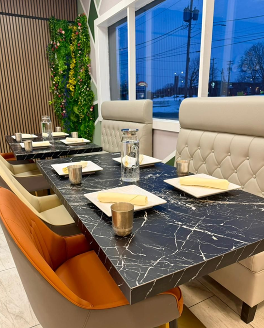 Restaurant tables set with water glasses, plates, and candles.  Orange and tan chairs. Marble-like tabletops.  Window with night view.