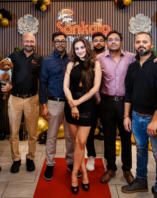 Group poses for a photo at an event: woman in black dress, men in various outfits, red carpet.