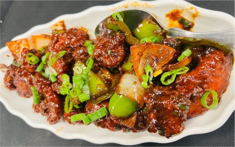Spicy, saucy chicken dish with green bell peppers, onions, and scallions in a white scalloped dish.