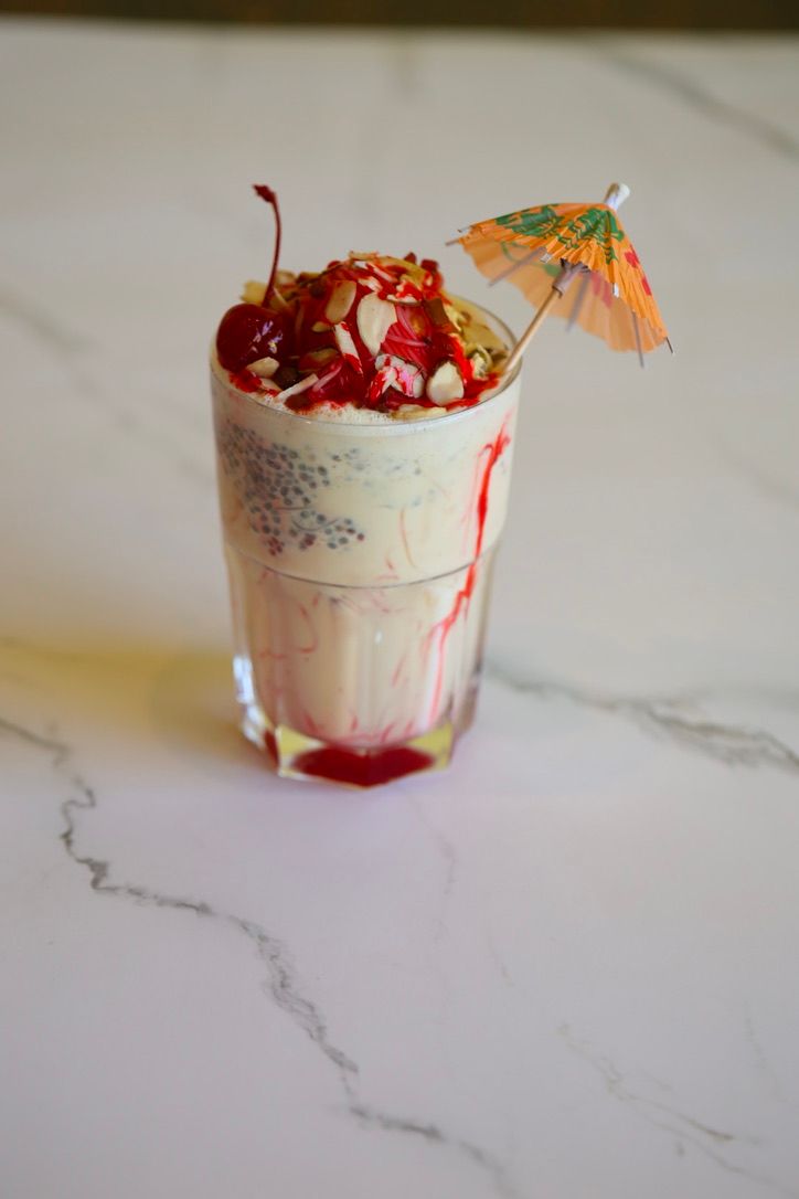 Tall glass of dessert with red syrup, topped with nuts, a cherry, and an umbrella, on a marble surface.