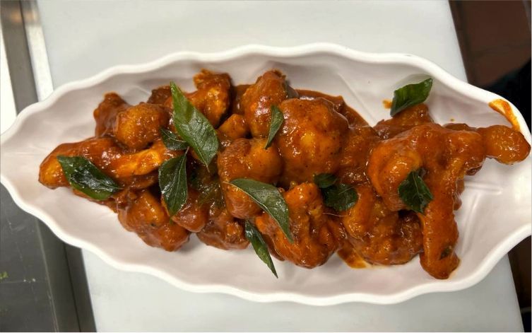 Fried cauliflower florets in red sauce, garnished with green curry leaves, served on a white leaf-shaped dish.