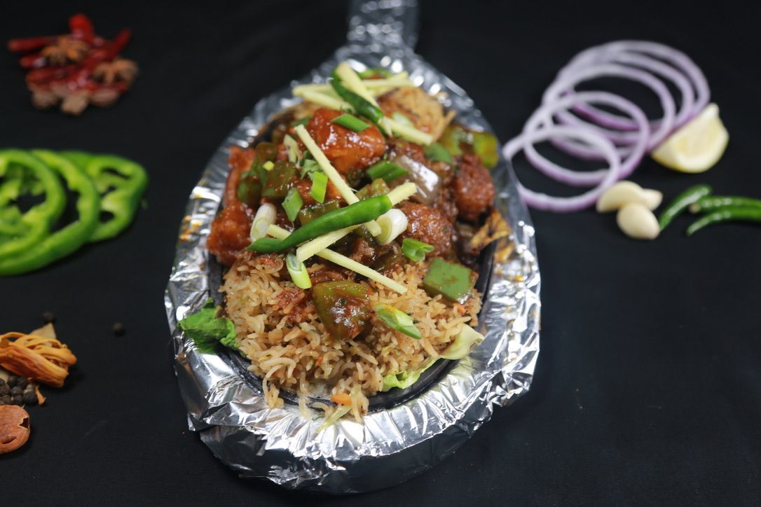 Sizzling plate of food with rice, vegetables, and garnish, on a black surface.