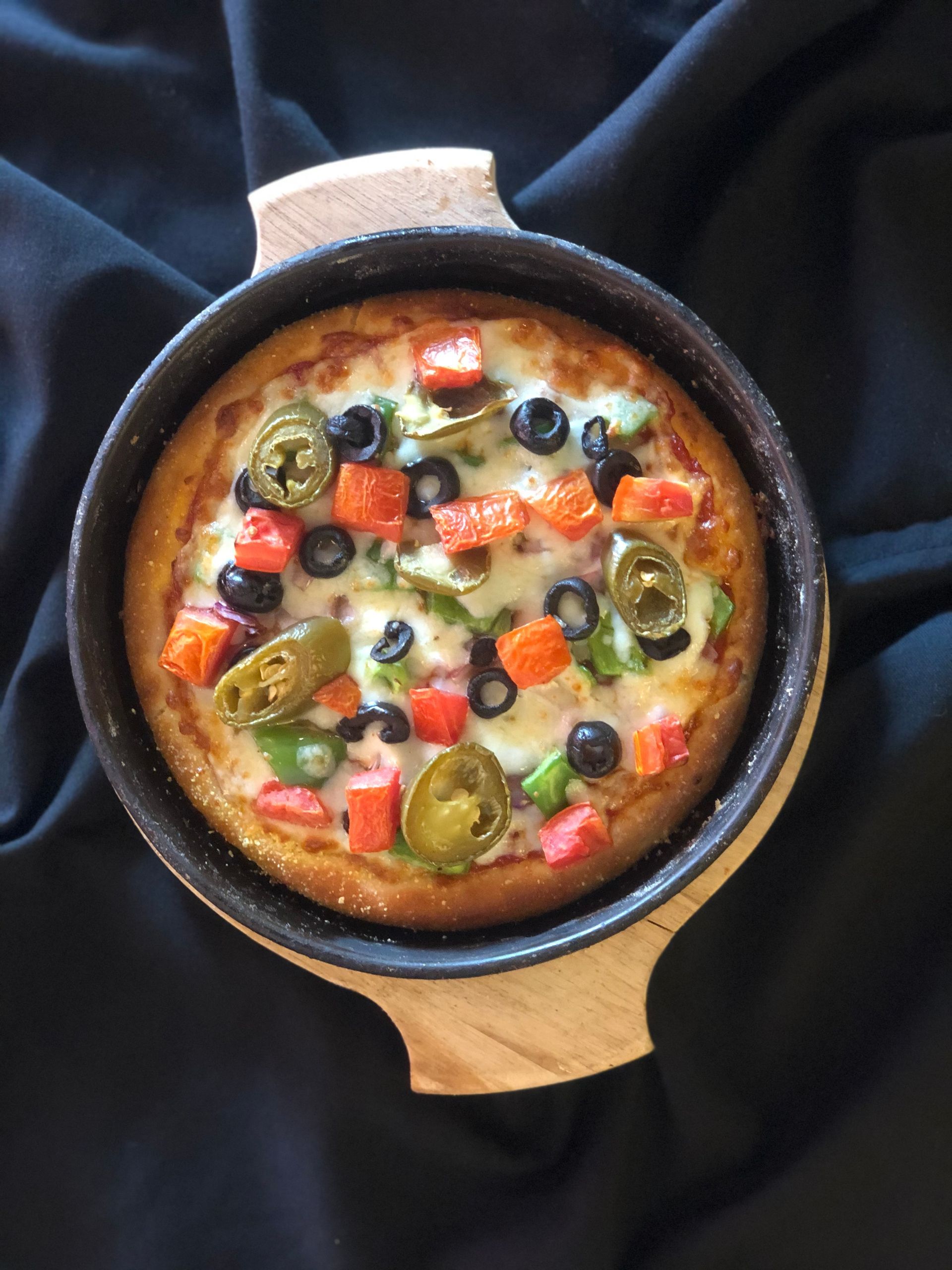 Pizza with olives, peppers, and cheese in a pan, set on a wooden base, against a black background.