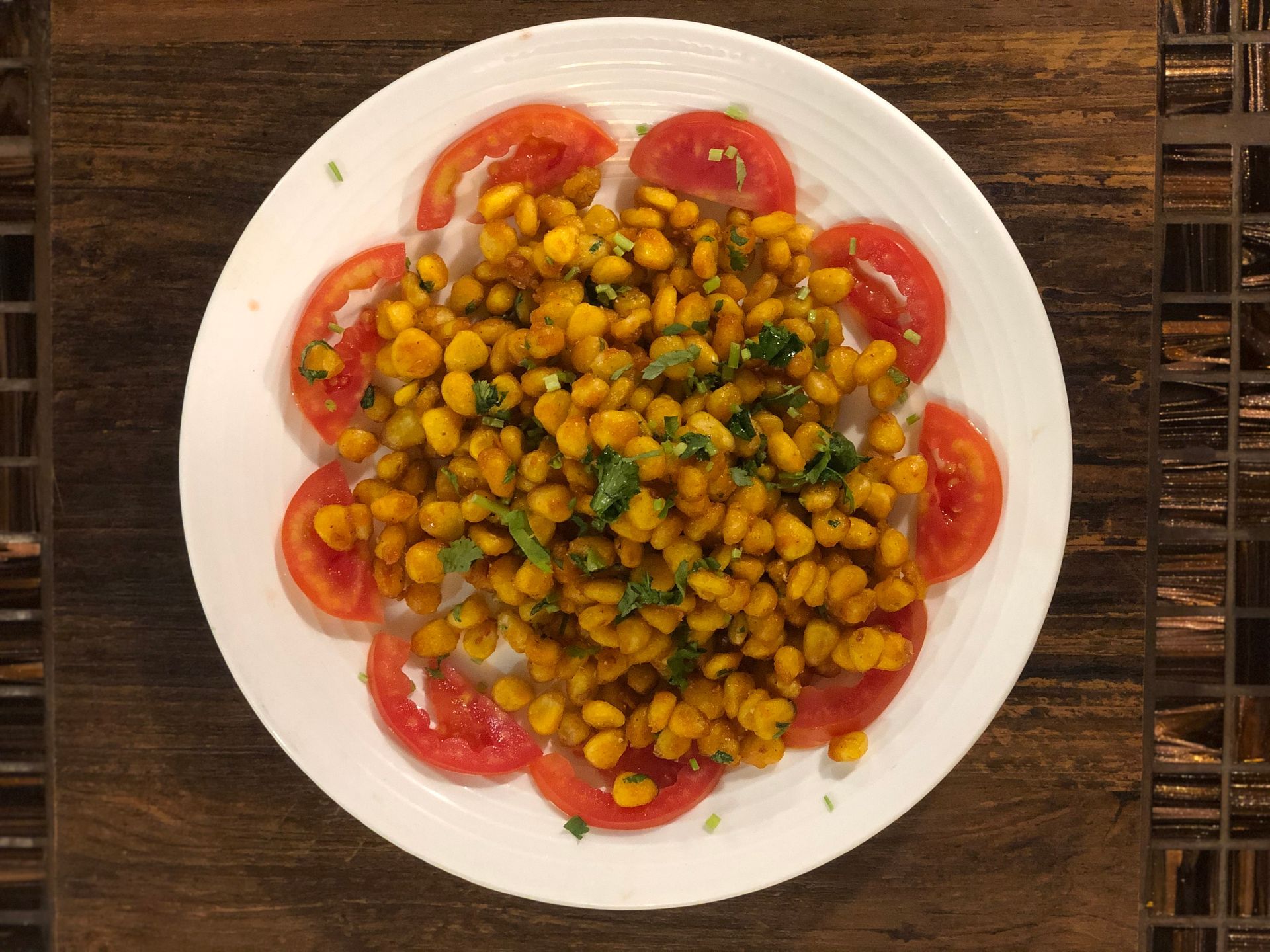 Corn dish with tomato slices on a white plate, on a brown tile surface.
