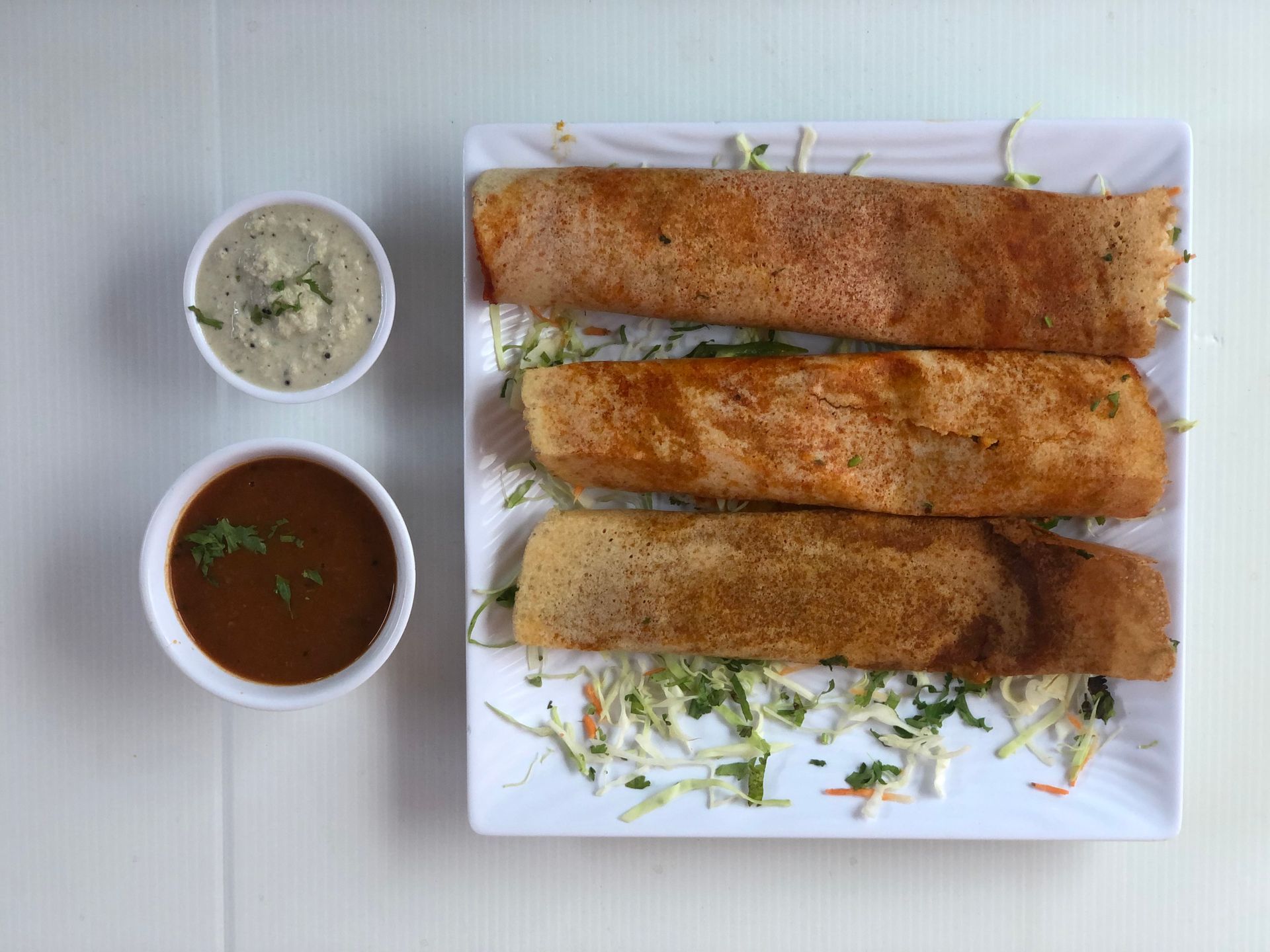 Three rolled dosas on a white plate with two small bowls of dipping sauces.