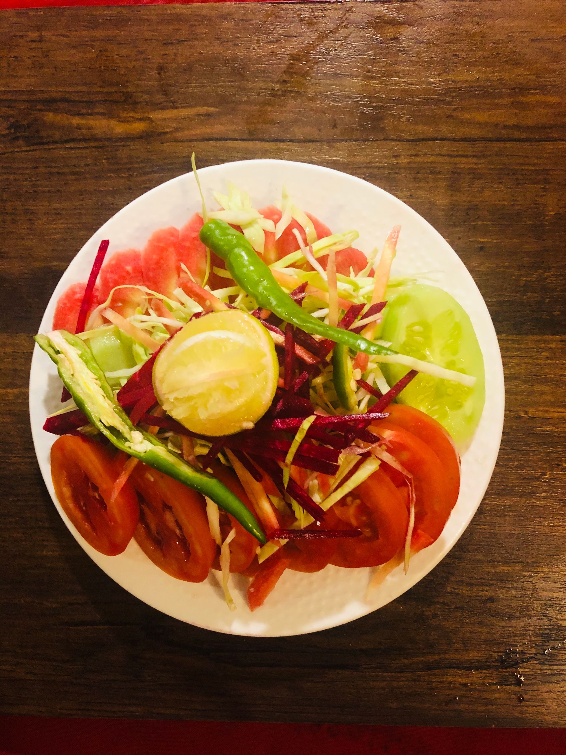 Salad on a white plate: tomatoes, cucumber, shredded cabbage, lime, and chili peppers on a wooden table.