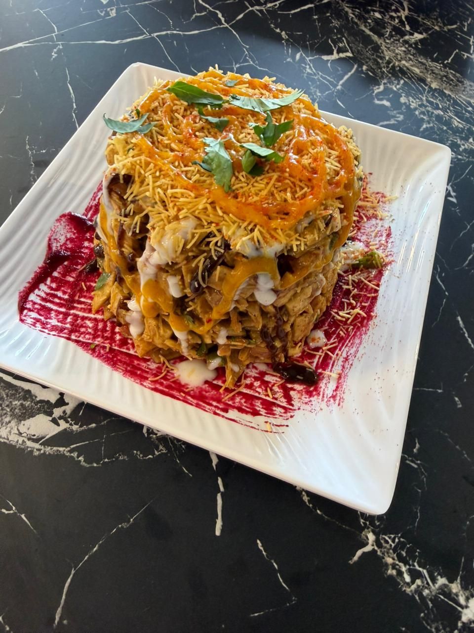 Tower of layered Indian chaat on a white plate, with beet puree and garnished with cilantro and crispy noodles.