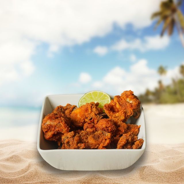 Fried chicken in a white bowl, topped with a lime slice, set on a sandy beach with a blurry ocean backdrop.