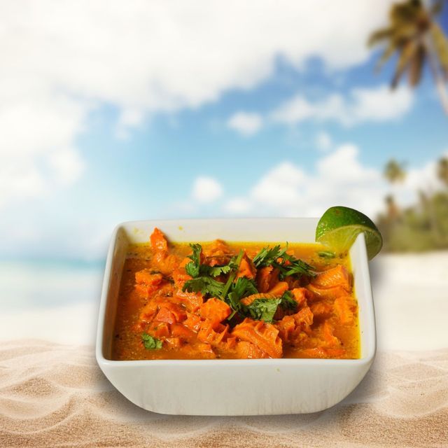 A white bowl of orange curry with cilantro and lime wedge on a sandy beach with a tropical background.