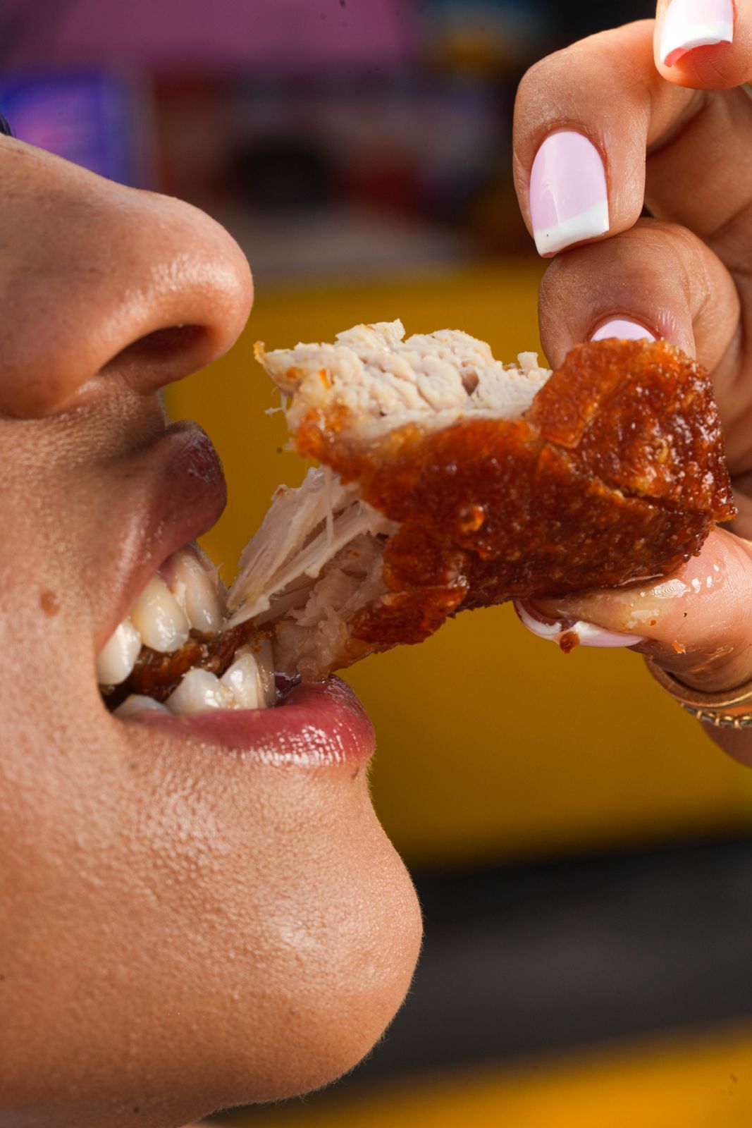 Person taking a bite of fried chicken wing, showing mouth, teeth, and hand holding the wing.