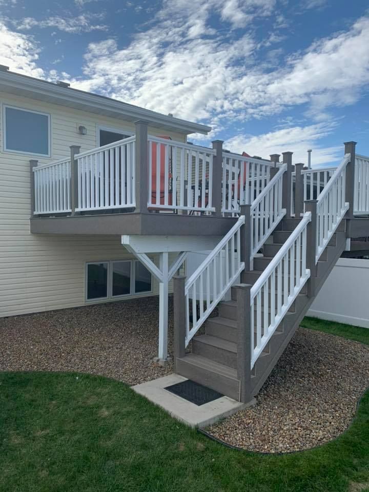 Beige house with a grey deck, white railing, and stairs; stone and grass in the yard.