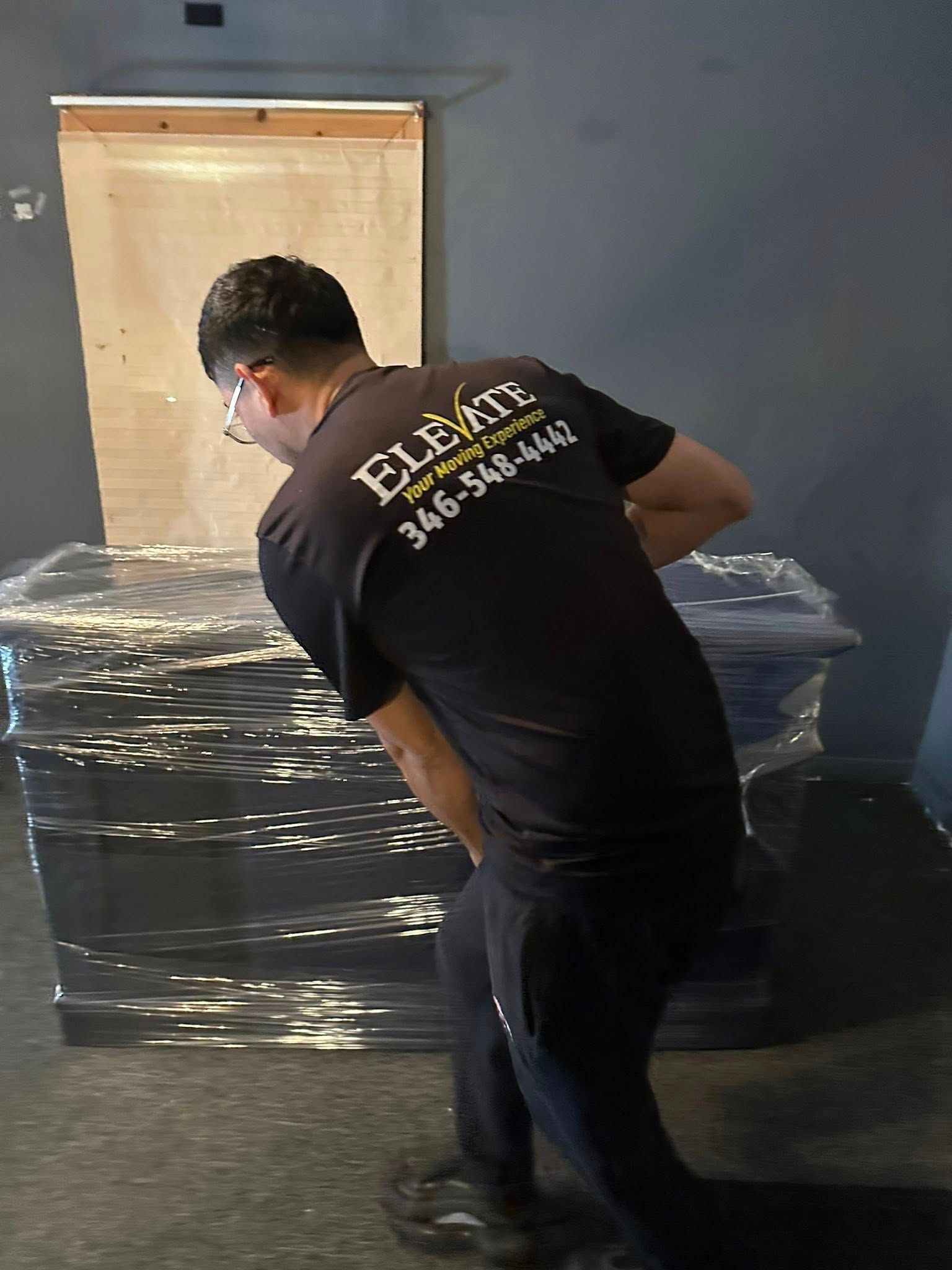 Man in black shirt with logo, lifting wrapped items. Black pants, indoor setting.
