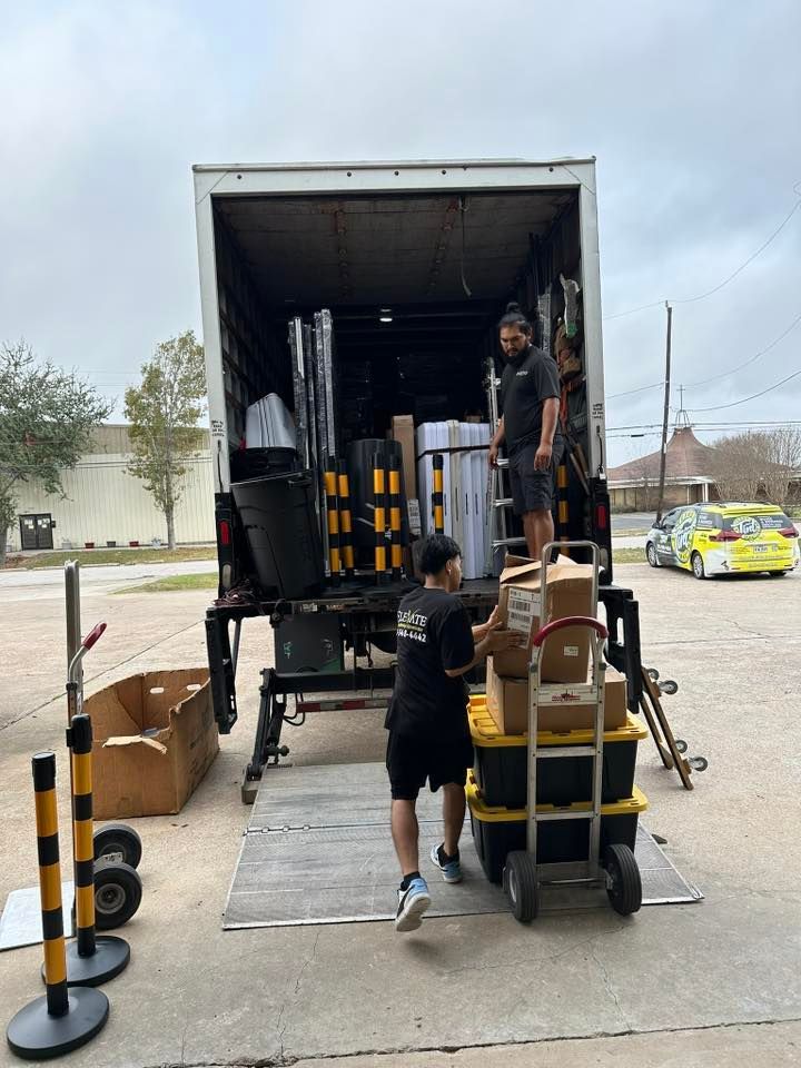 Two people loading boxes onto a truck with a ramp outdoors. Yellow and black caution poles are nearby.