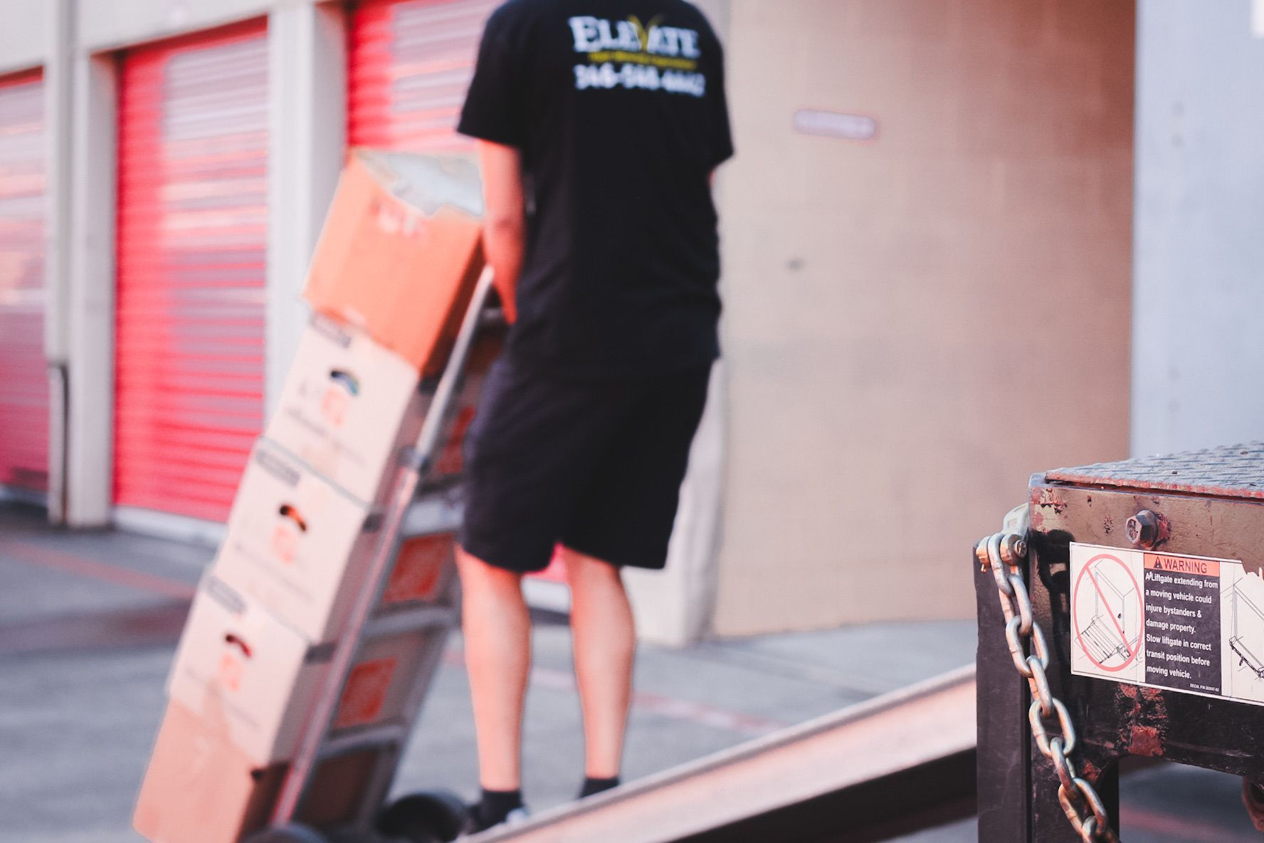 Person in black shirt and shorts carrying stacked moving boxes up a ramp into a storage unit.