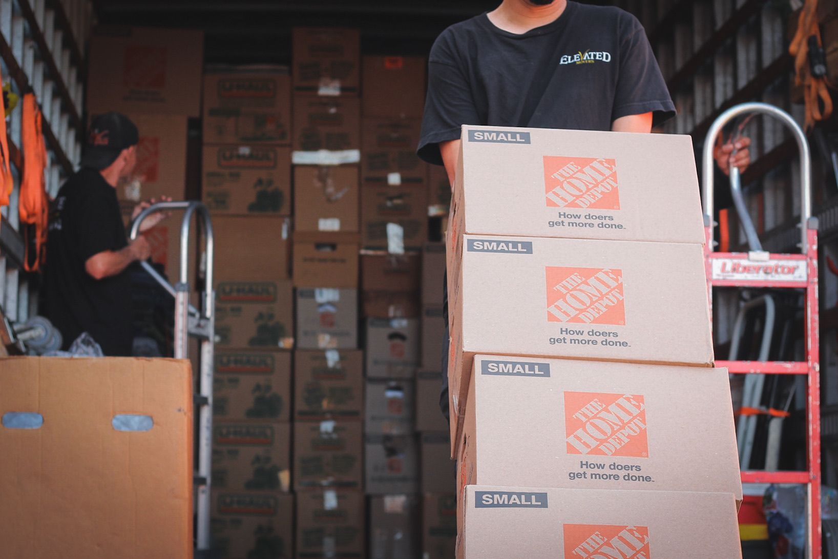 Movers loading moving boxes into a truck, one person carrying stack of boxes, one using a dolly.