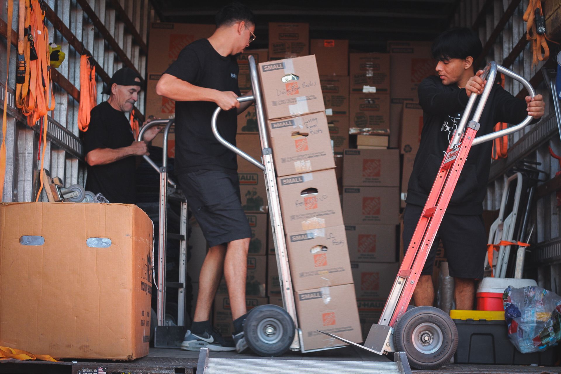 Three people loading a moving truck with boxes. Two use hand trucks, one is inside the truck. Boxes are stacked.