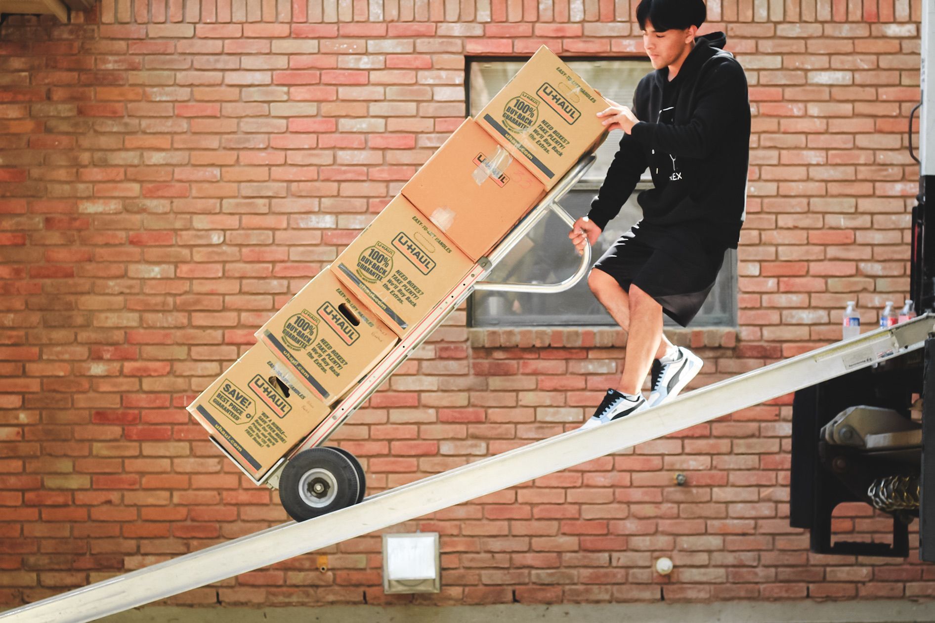 Man pushes boxes on a hand truck up a ramp; against a brick wall.