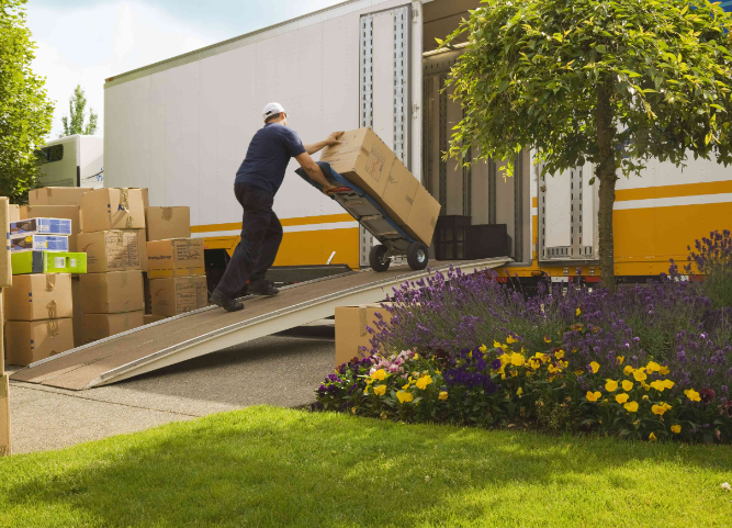 Mover loading boxes into a truck via a ramp outside a house with flowers and trees.