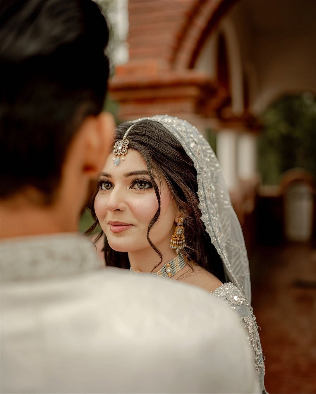 A person in a decorative white bridal veil and jewelry gazes softly towards the camera, partially framed by another person.