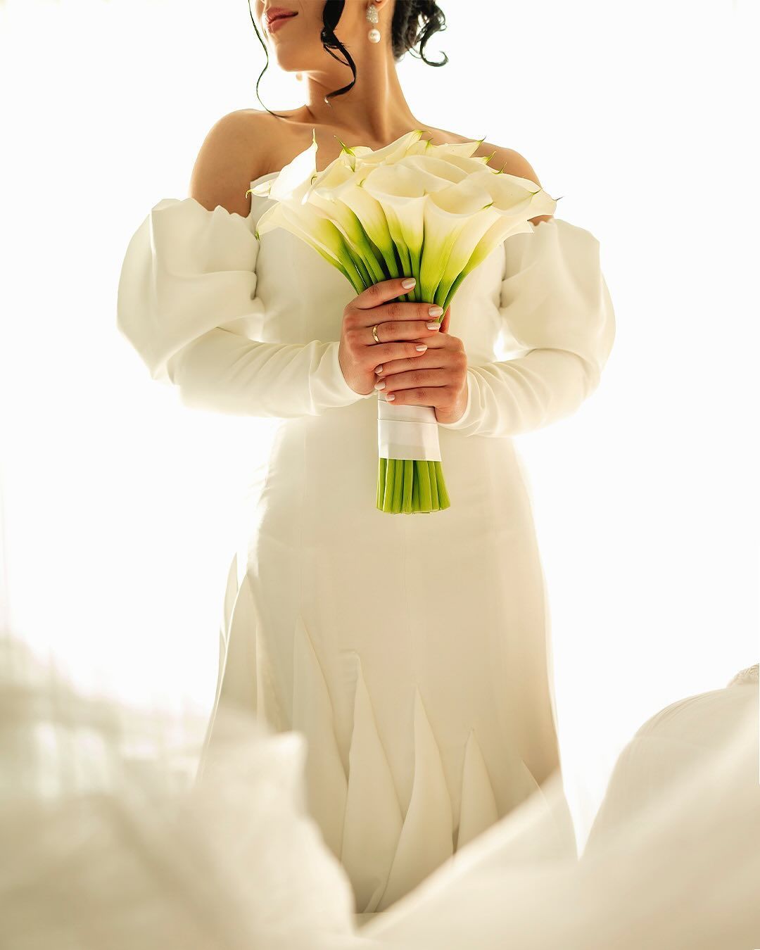 A person in an off-the-shoulder white wedding gown holds a bouquet of white calla lilies against a bright background.