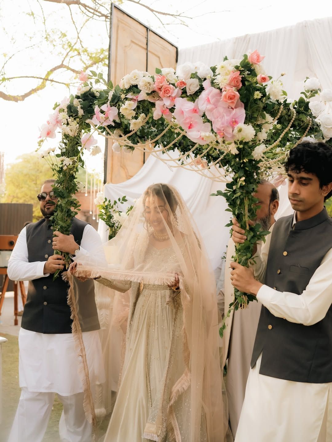 A bride walks under a floral arch held by two attendants during a wedding ceremony.