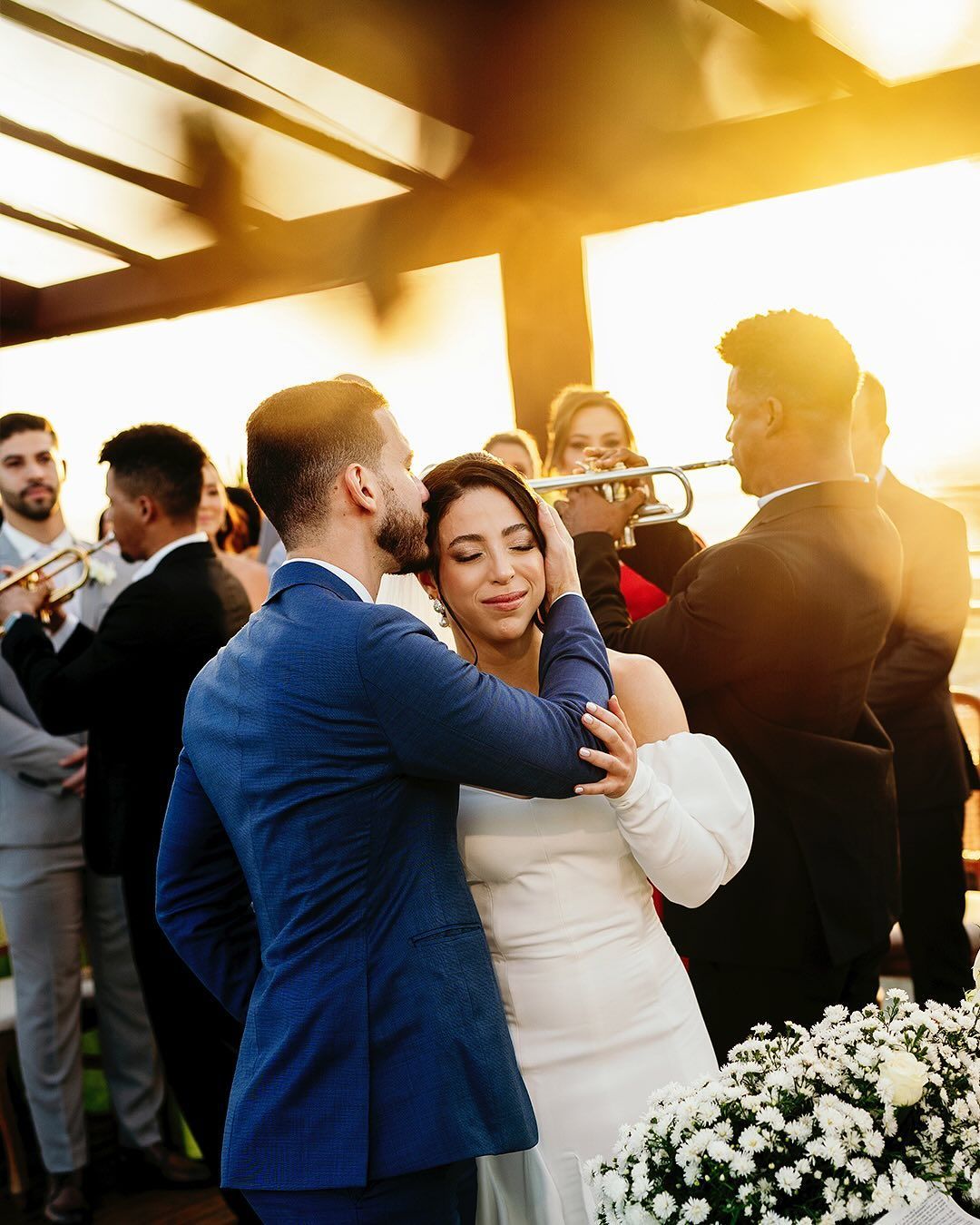 A couple embraces at a sunset wedding while a musician plays a trumpet in the background.