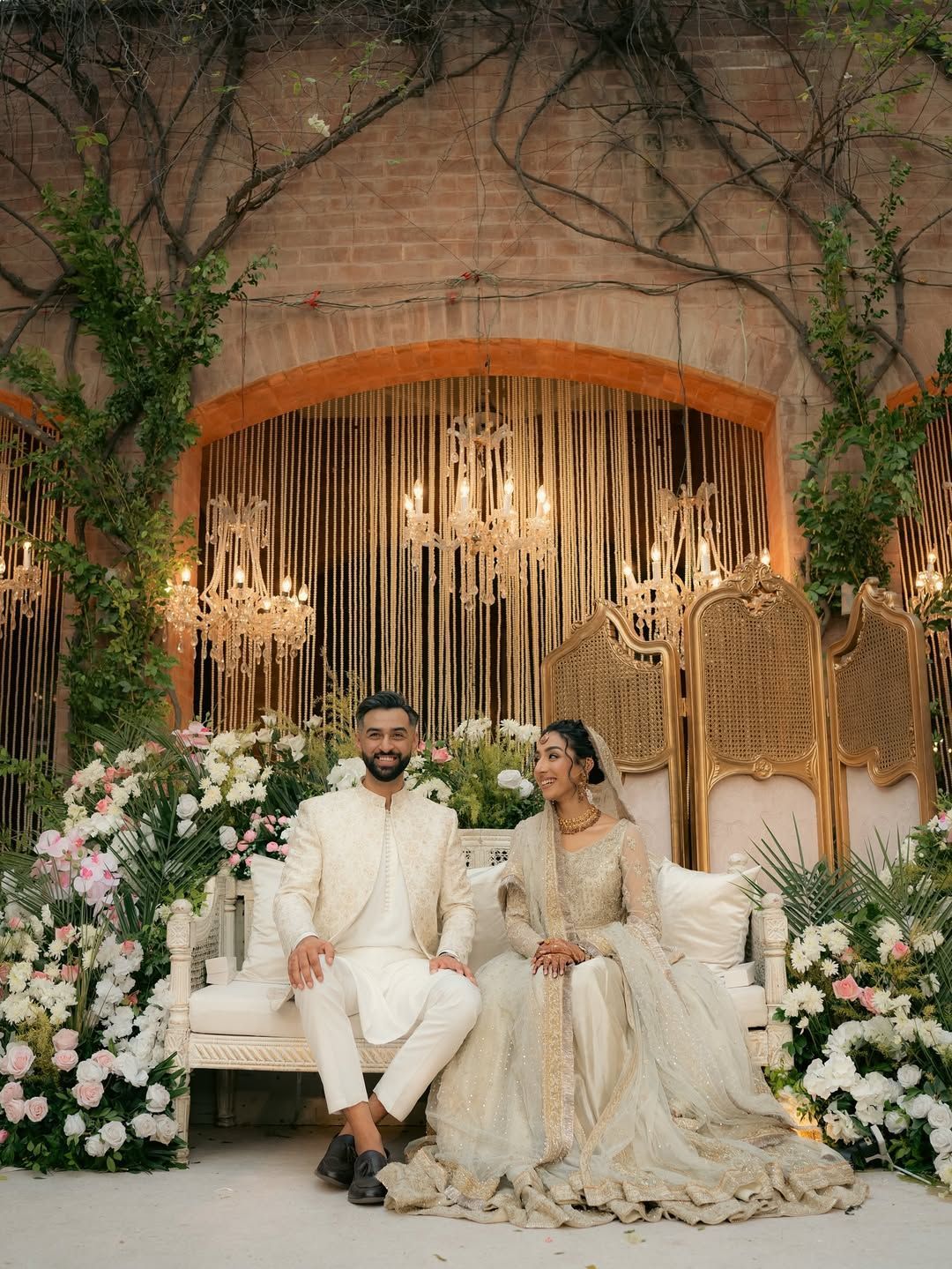 A couple in traditional cream-colored formal attire sits on a white bench, surrounded by floral arrangements and lanterns.