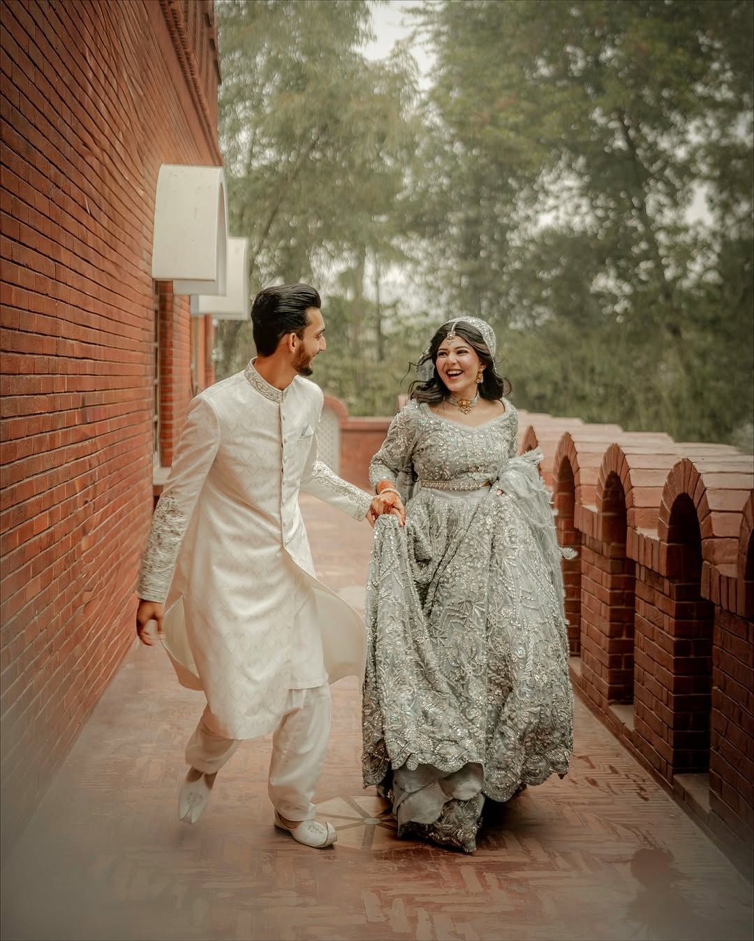 A joyful couple in traditional wedding attire walks hand-in-hand along a brick walkway outdoors.