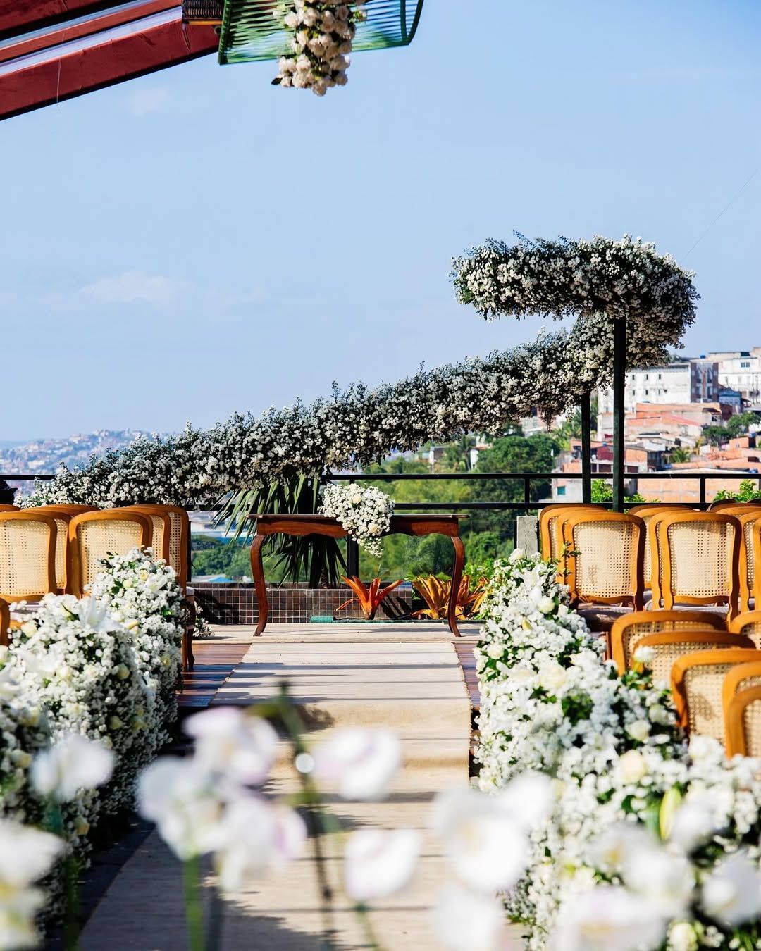 Wedding aisle with white floral arrangements, wooden chairs, and a sweeping overhead greenery arch against a cityscape.
