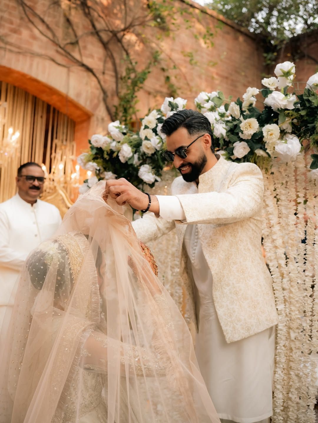 A person in a cream-colored sherwani places a veil over a partner's head during an outdoor wedding ceremony.