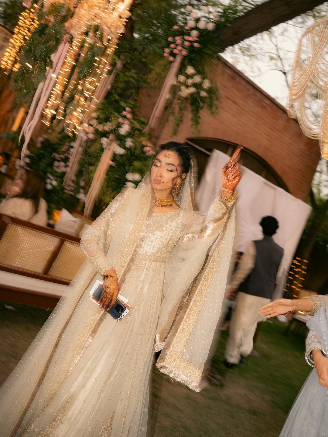 A person in an ornate, cream-colored formal gown and head covering posing gracefully at an outdoor event with warm lighting.