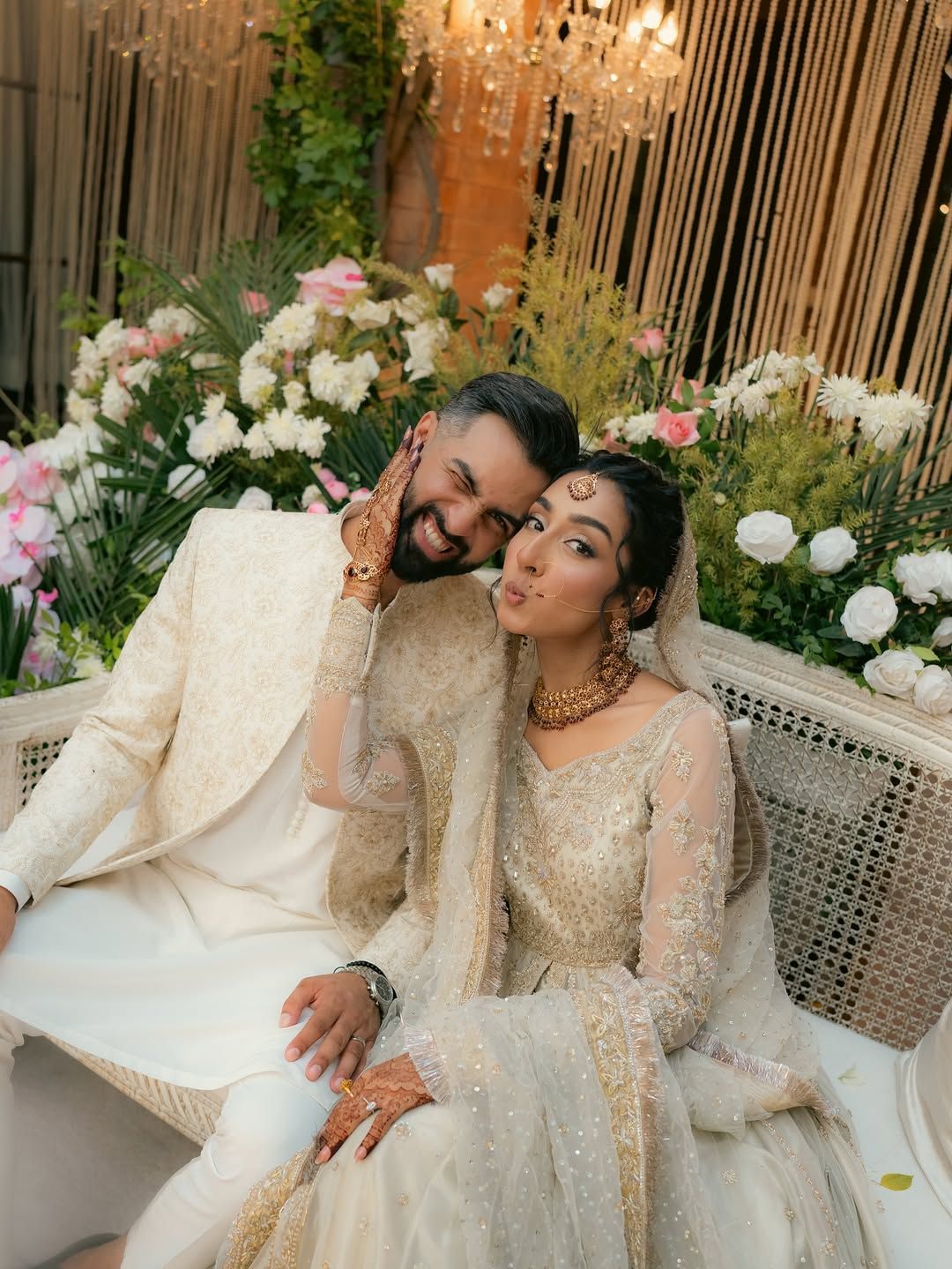 A bride and groom in traditional cream-colored wedding attire sitting together, posed affectionately against flowers.